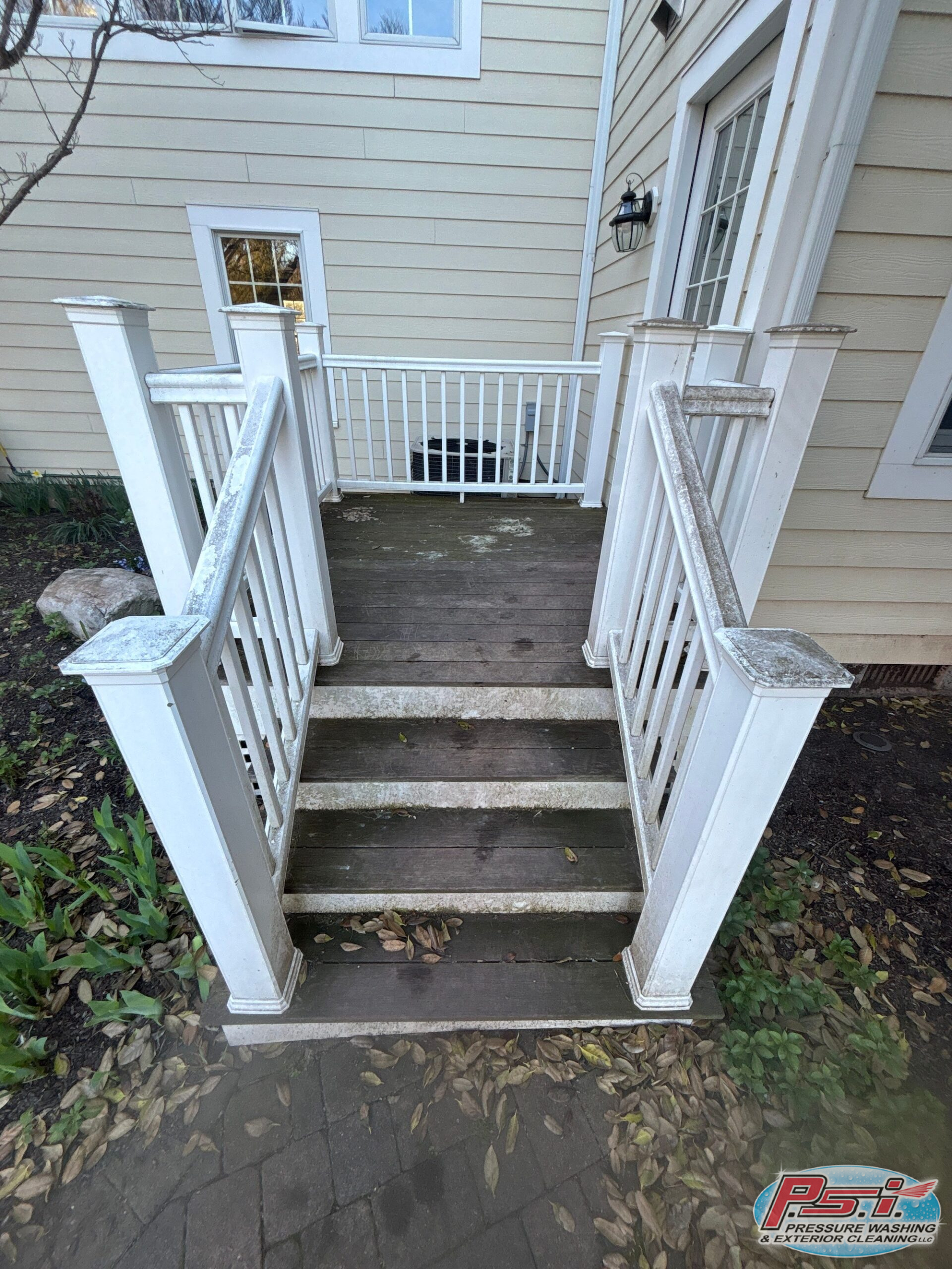 Exterior wooden staircase with white railings and posts leading to a door with a window; leaves scattered on a brick pathway.