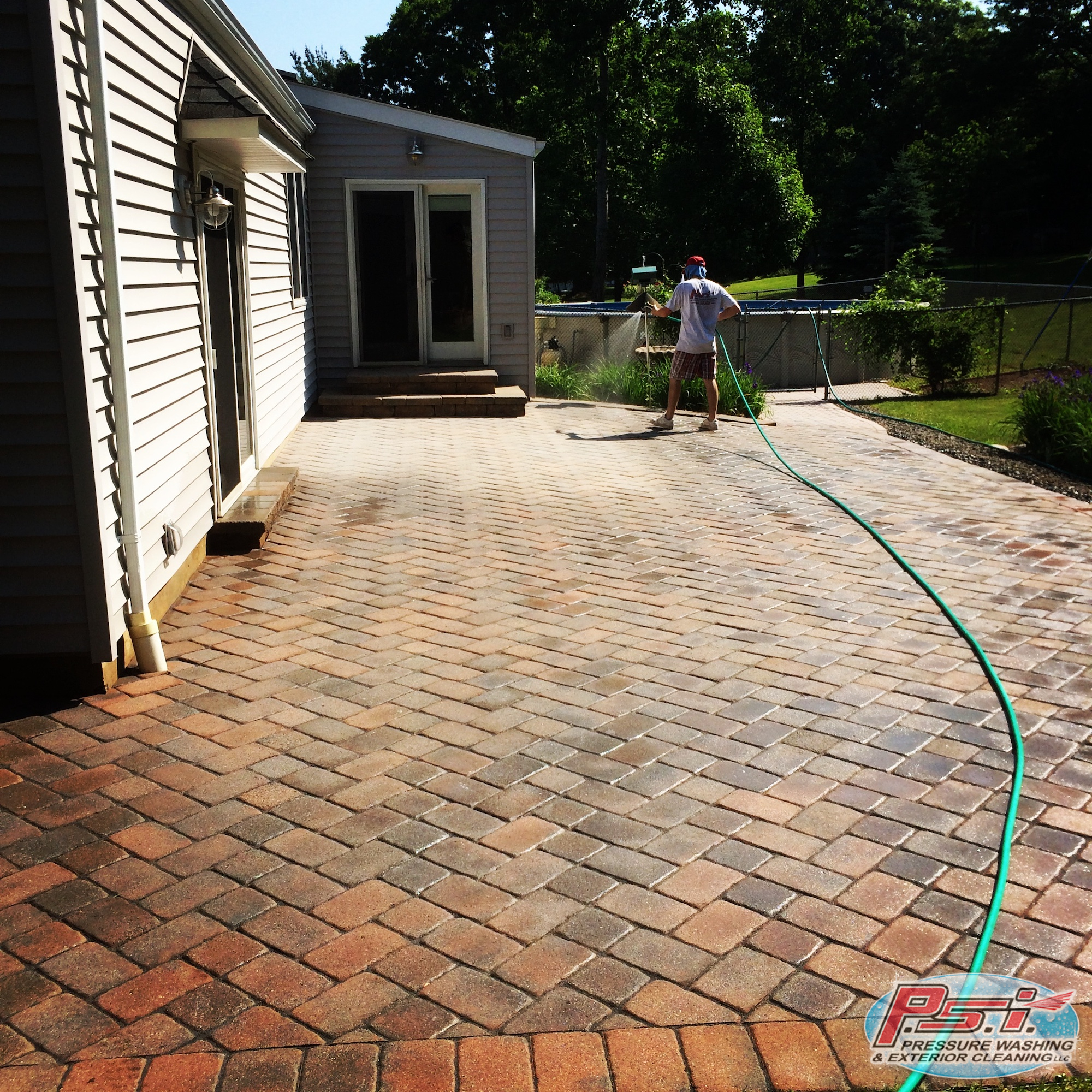 Brick-paved patio beside a light-colored house with sliding door; man in shorts using a long green hose, likely cleaning exterior area.