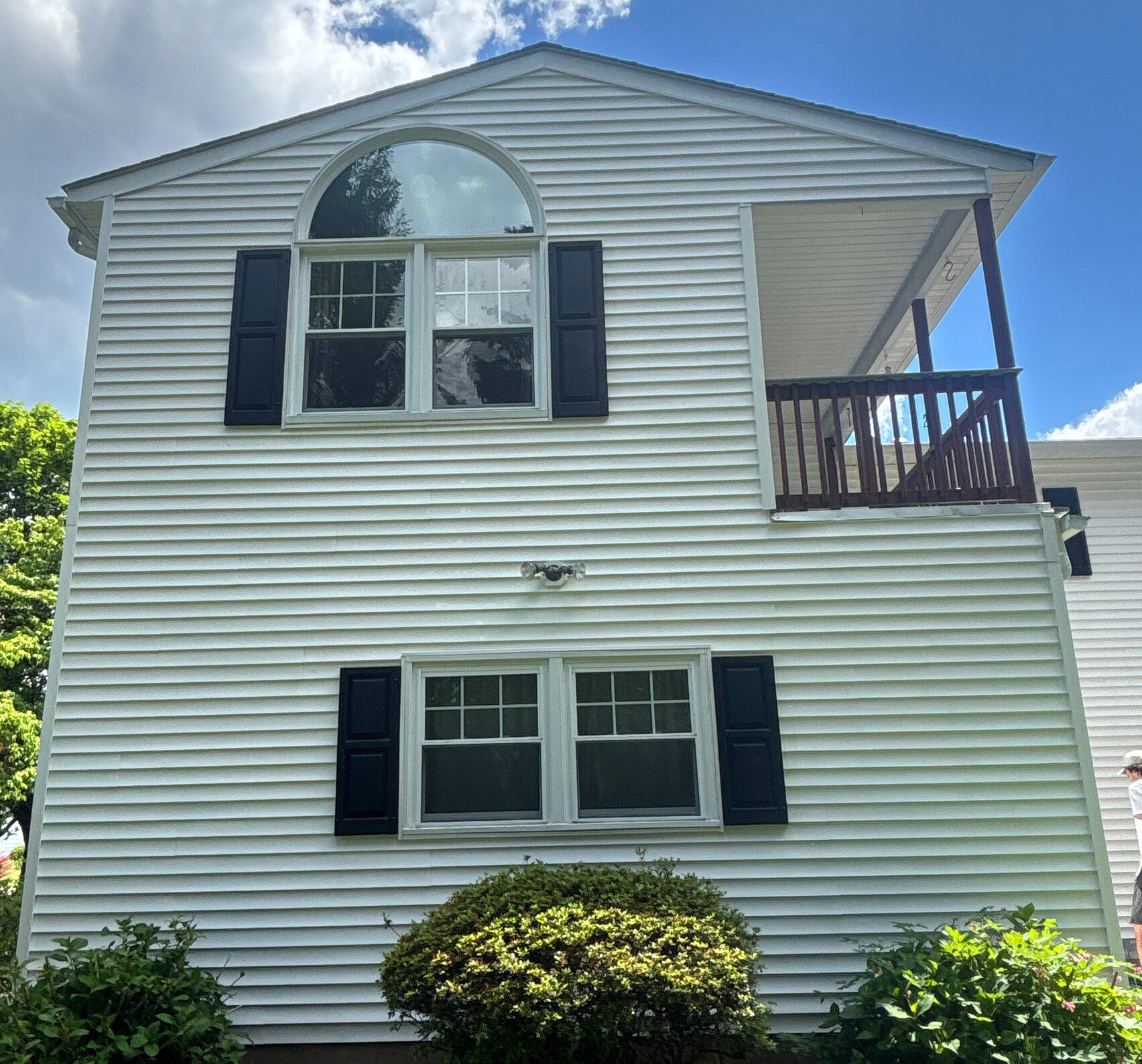White two-story house with an arched second-floor window framed by black shutters and a wooden balcony on the right; landscaped front area.