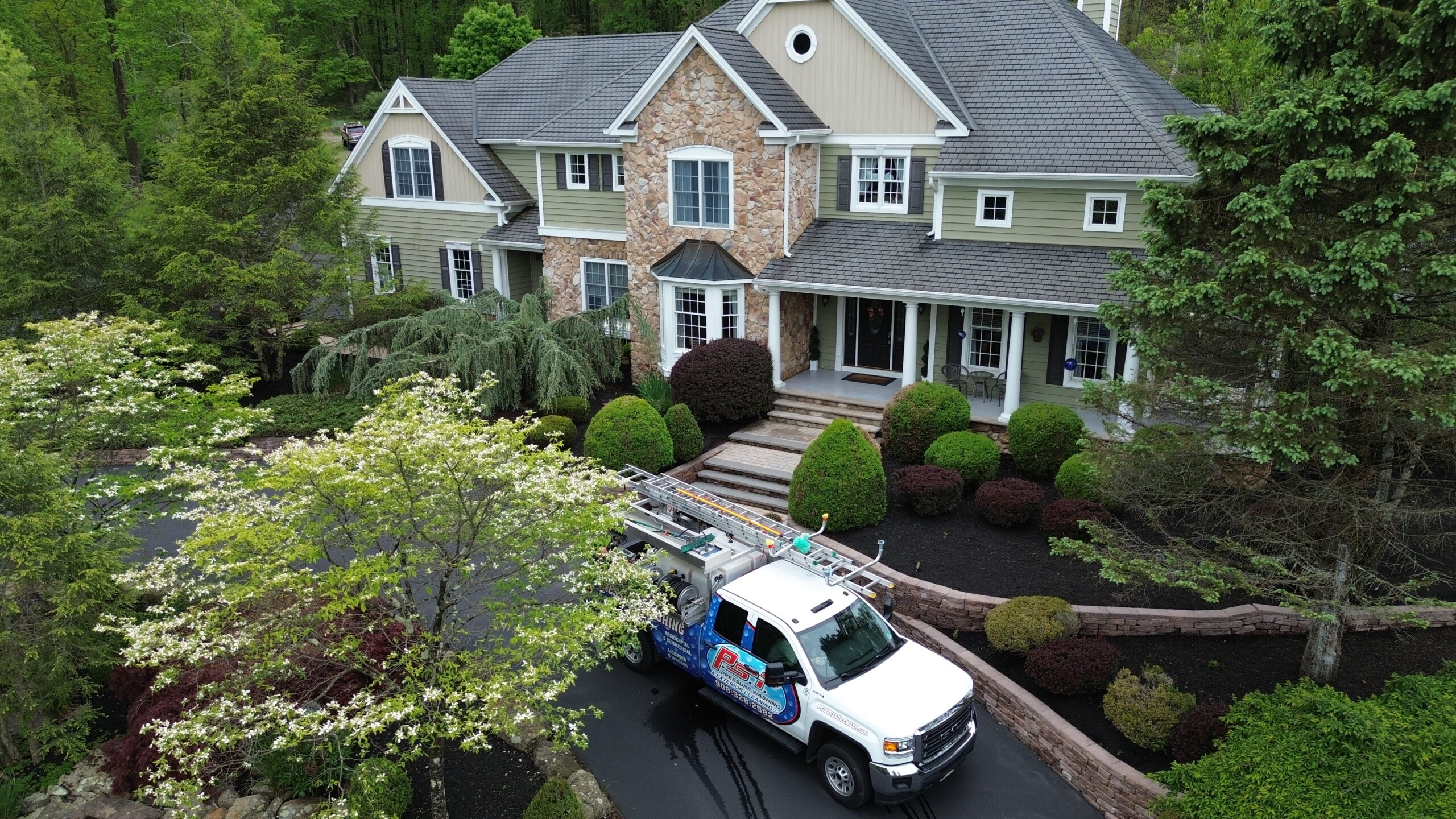 Front view of a large stone-and-siding house with manicured shrubs and a ladder-truck parked in a curved driveway between trees.