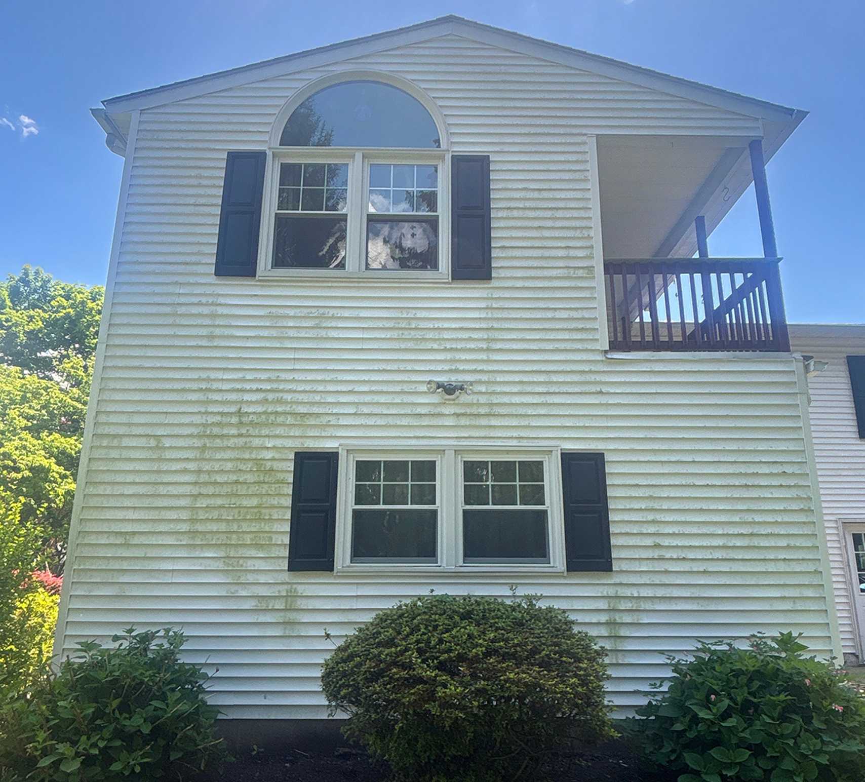 White two-story house with arched upstairs window, black shutters, and a right-side balcony, set against a blue sky with front shrubs in the foreground.