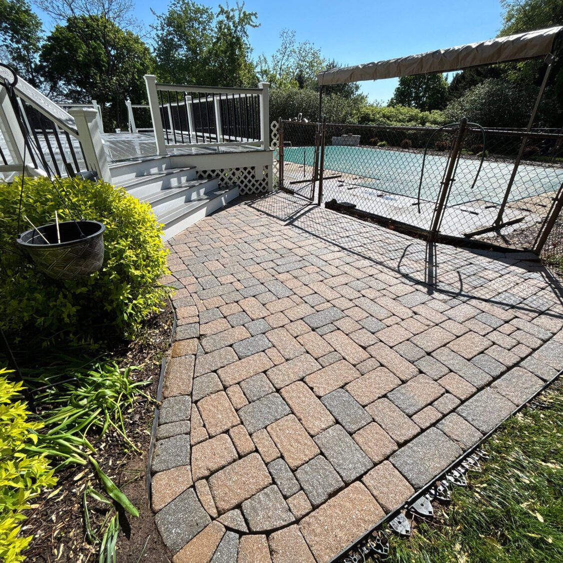 Curved brick patio beside a white-railed deck overlooking a backyard pool under a clear blue sky, with trees surrounding.