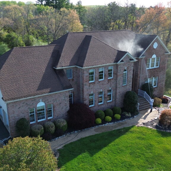Aerial view of a large brick house with smoke rising from the roof as firefighters work outside a white truck and ladder in the driveway and well kept landscaping surrounding the home