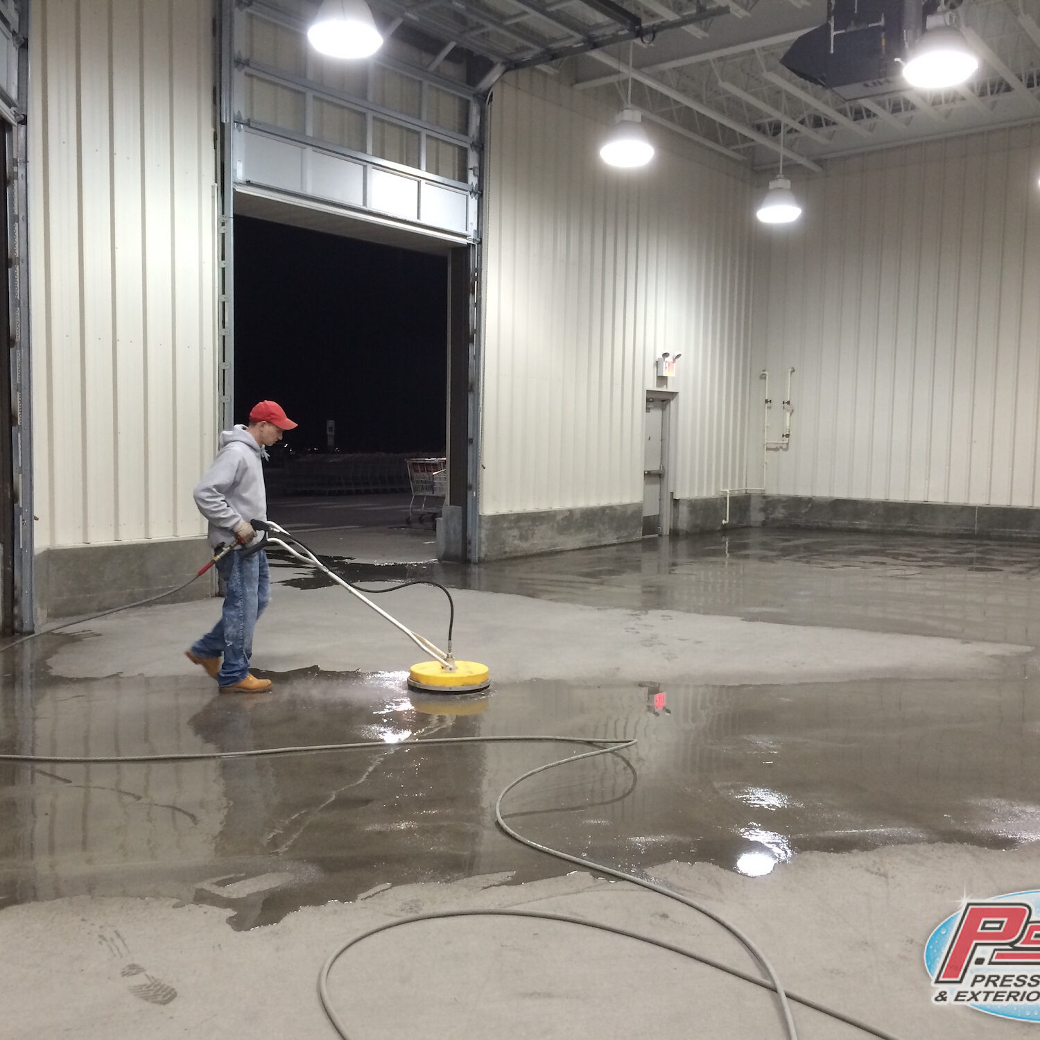 Worker in a red cap and gray hoodie operates a circular floor cleaning machine inside a large industrial bay; wet concrete floor.