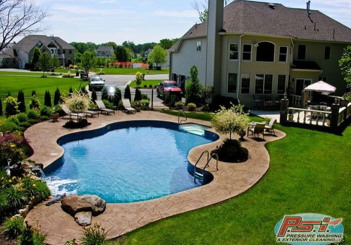 The backyard features a kidney-shaped swimming pool surrounded by a stone patio. Lounge chairs are placed around the pool, highlighting the pristine pool area cleaning. A well-manicured lawn and garden plants border the pool area, with a large house featuring an outdoor deck and a red car in the driveway in the background.