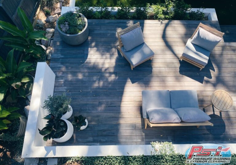 Aerial view of a clean deck featuring a wooden patio with cushioned chairs and a loveseat. The patio, perfect for the spring season, is adorned with potted plants and bathed in a mix of sunlight and shadow. Surrounding the patio is greenery, adding a natural ambiance to the cozy outdoor space.