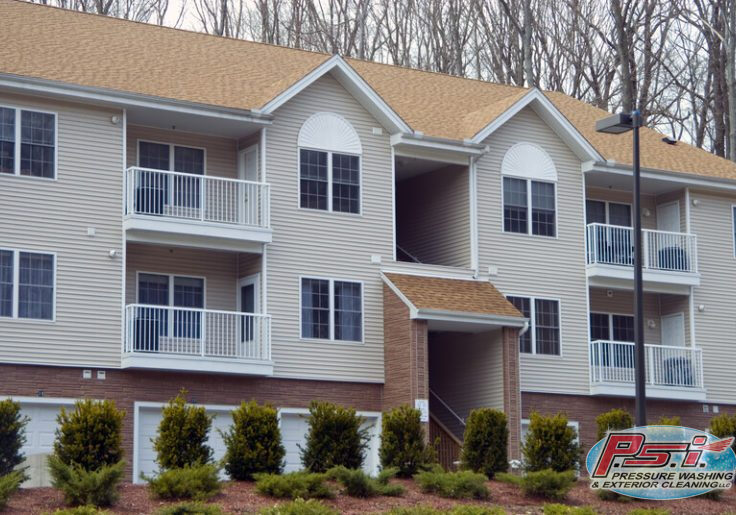 A two-story apartment building with beige siding and a brown roof features balconies with white railings. Bushes and small trees are planted in front of the building, and a streetlight is visible on the right side. The design allows for easy apartment cleaning, ensuring every unit remains pristine.