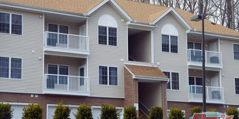 A two-story apartment building with beige siding and a brown roof features balconies with white railings. Bushes and small trees are planted in front of the building, and a streetlight is visible on the right side. The design allows for easy apartment cleaning, ensuring every unit remains pristine.