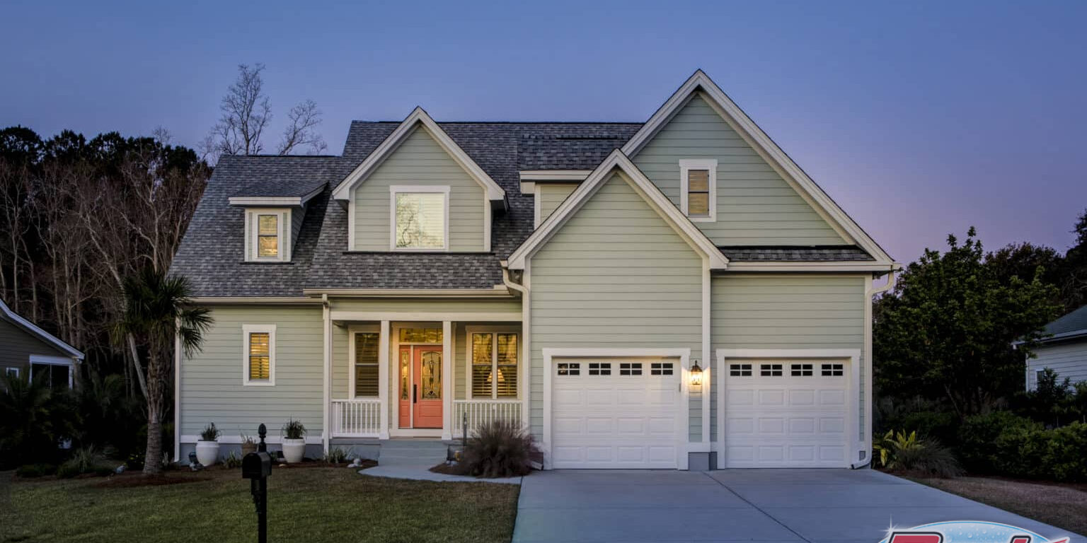Two-story house with light green, clean siding and a two-car garage at dusk. The home boasts multiple windows, a well-maintained lawn, and significant curb appeal with its lit front porch featuring columns. A driveway leads to the garage doors, and trees are visible in the background.