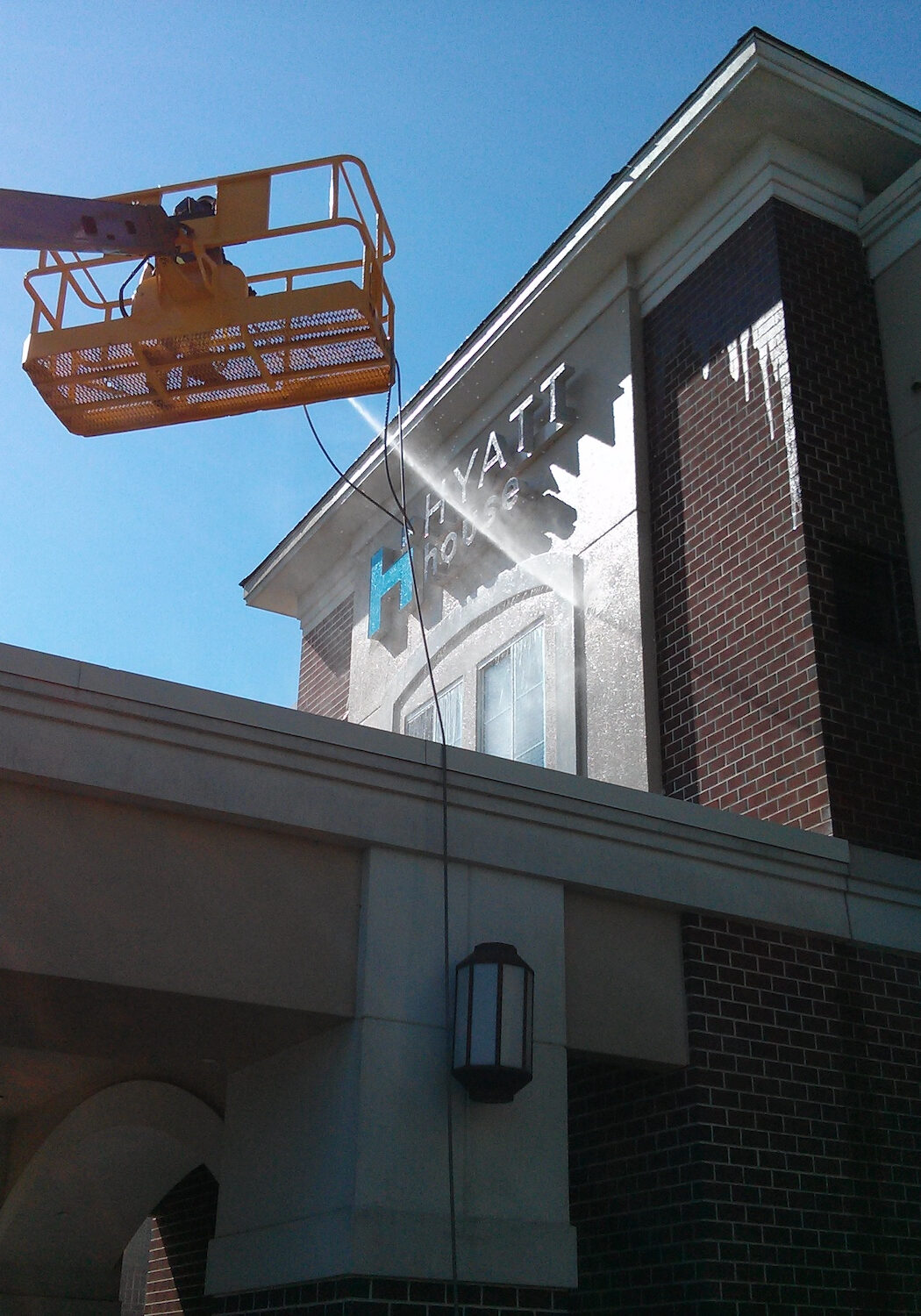Orange elevated work platform (bucket lift) extended toward a brick building on a sunny day, against a blue sky.