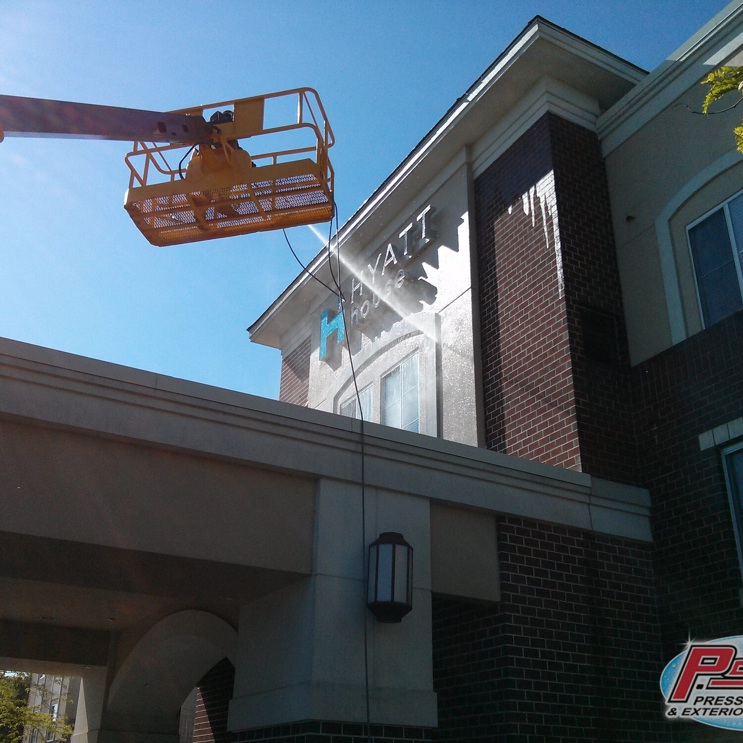 Orange elevated work platform (bucket lift) extended toward a brick building on a sunny day, against a blue sky.