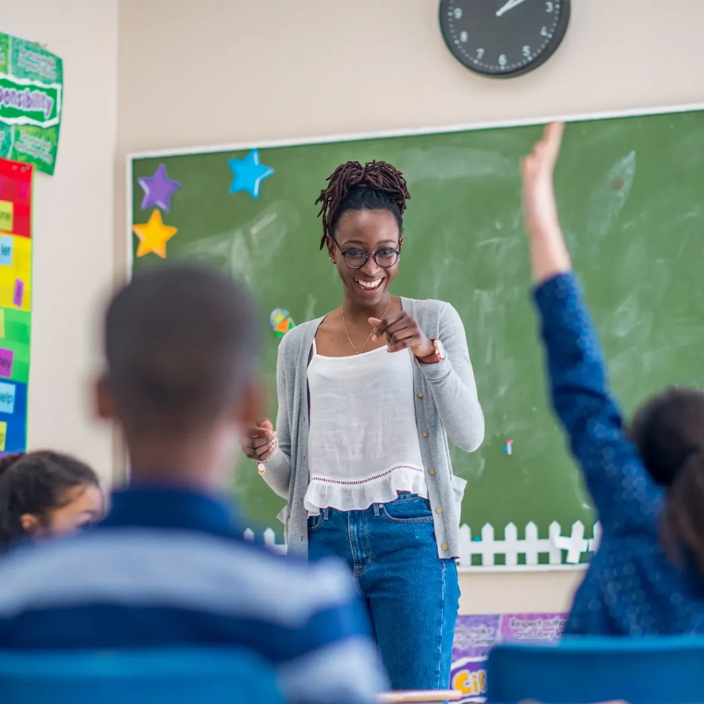 Teacher smiling and pointing at students in a classroom with a green chalkboard and colorful wall posters, engaging the class.