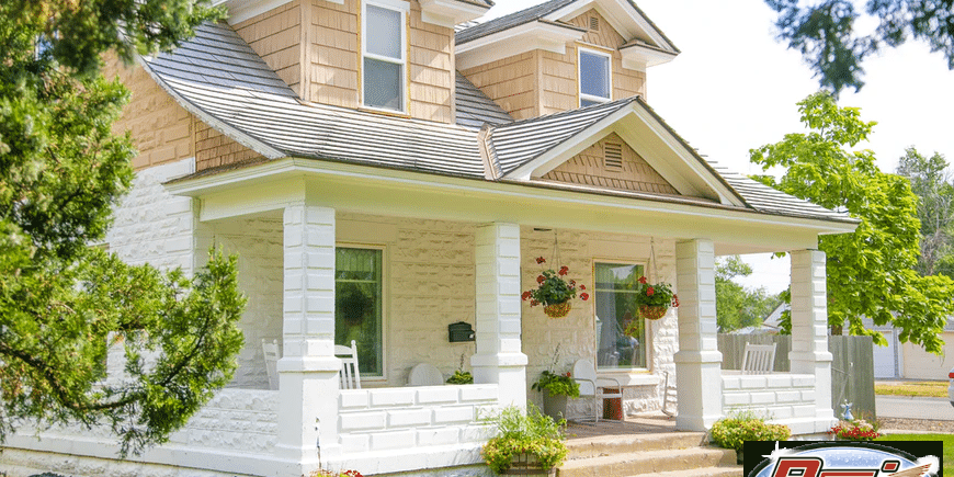 A charming white house with a cozy porch adorned with hanging flower baskets and potted plants. The home's exterior features dormer windows on the upper level and a well-maintained lawn, revived each spring. Tall trees frame the scene, adding to the peaceful, inviting atmosphere.