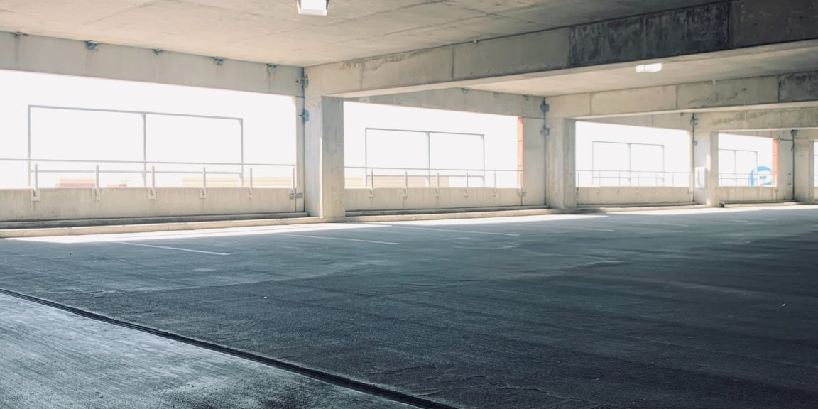 A well-lit, clean parking garage with concrete floors and columns. Large windows on the sides allow natural light to flood in. The area is spacious, with visible parking lines on the ground, highlighting the excellent garage cleanliness and maintenance.