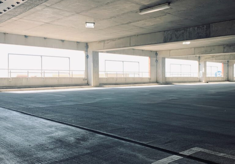 A well-lit, clean parking garage with concrete floors and columns. Large windows on the sides allow natural light to flood in. The area is spacious, with visible parking lines on the ground, highlighting the excellent garage cleanliness and maintenance.