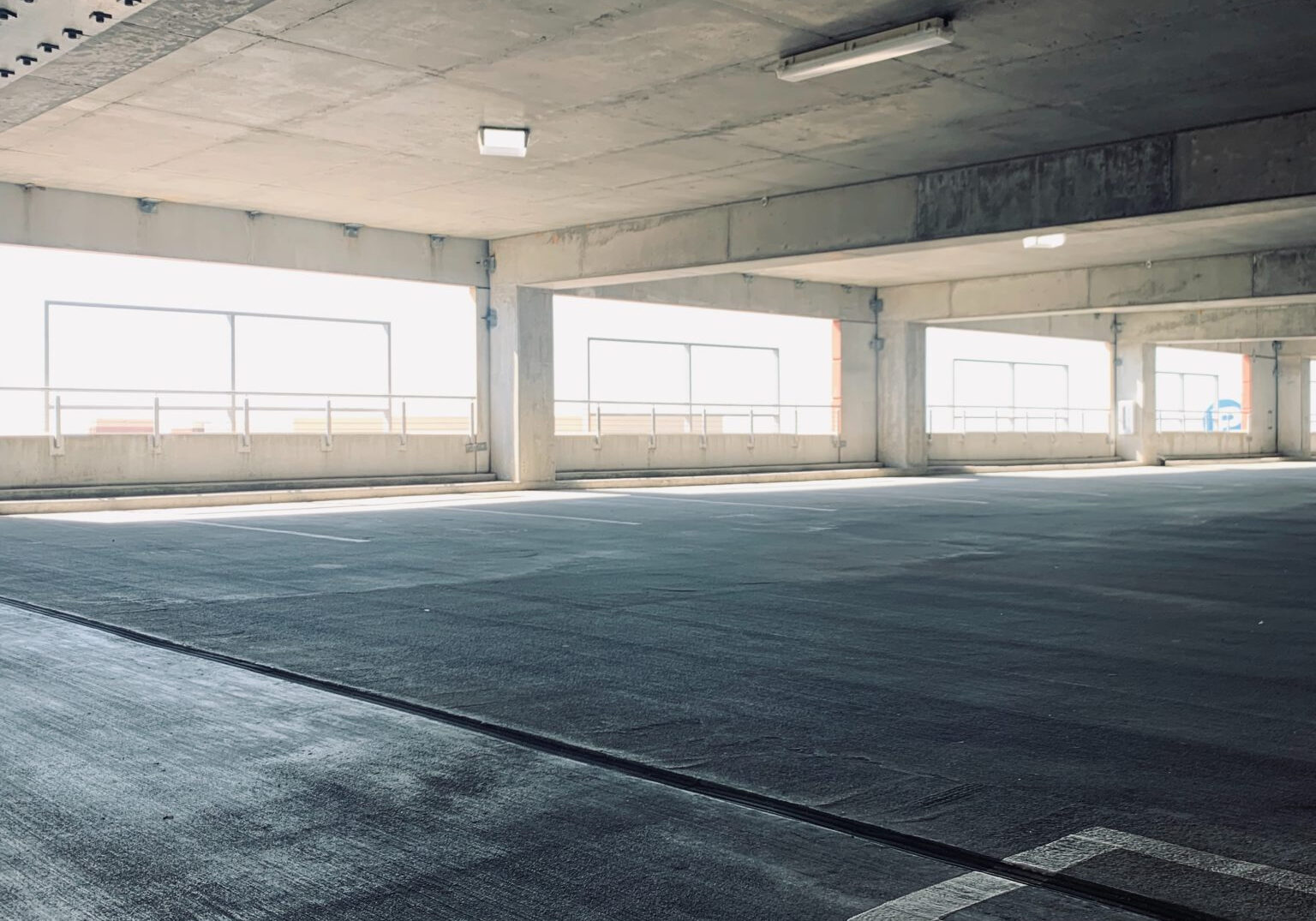 A well-lit, clean parking garage with concrete floors and columns. Large windows on the sides allow natural light to flood in. The area is spacious, with visible parking lines on the ground, highlighting the excellent garage cleanliness and maintenance.