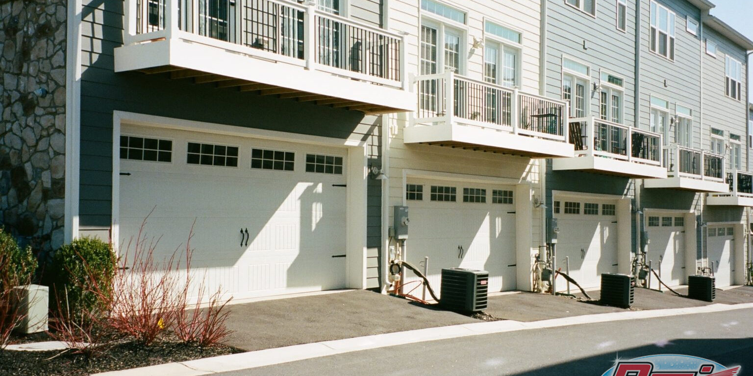 A row of modern townhomes with beige and gray exteriors. Each unit has a white garage door, balconies with railings above the garages, and air conditioning units beside the driveways. The street in front is clean and vacant, with minimal landscaping.