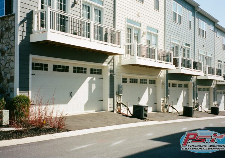 A row of modern townhomes with beige and gray exteriors. Each unit has a white garage door, balconies with railings above the garages, and air conditioning units beside the driveways. The street in front is clean and vacant, with minimal landscaping.