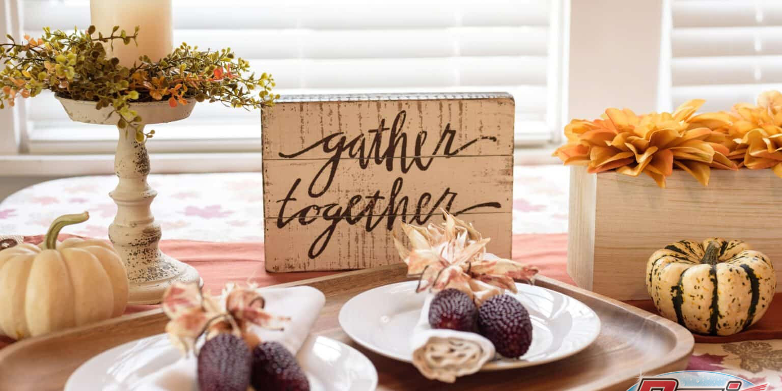 A cozy dining table setup for Thanksgiving featuring a "gather together" sign, decorative pumpkins, and dried corn on white plates. The table is adorned with a candle centerpiece and orange flowers, creating a warm, autumnal ambiance to help you prepare your home for the holiday season.